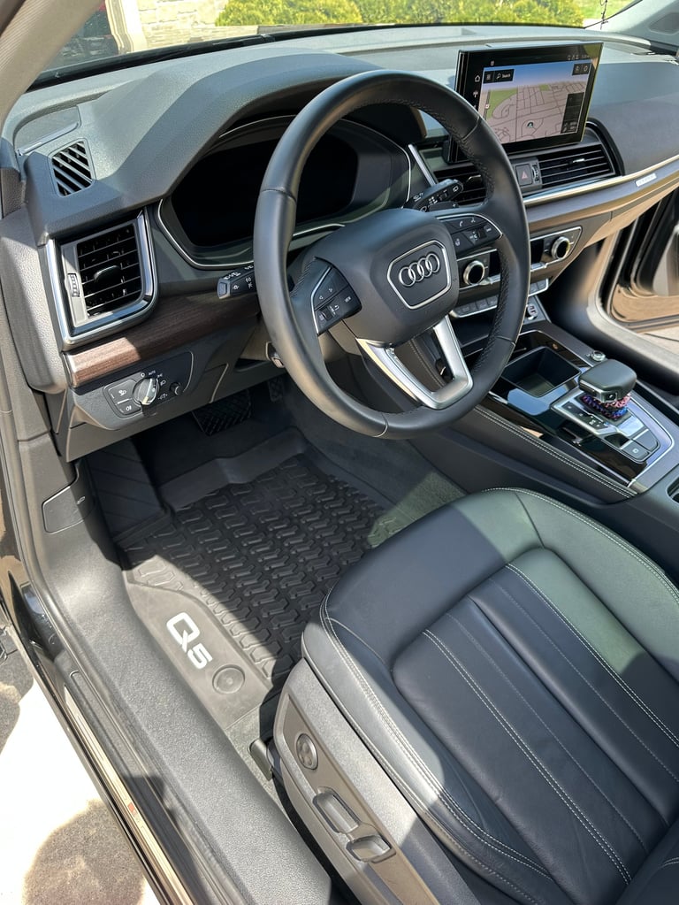 Interior of Audi vehicle showing steering wheel, dashboard, touchscreen display, and leather seats with floor mats